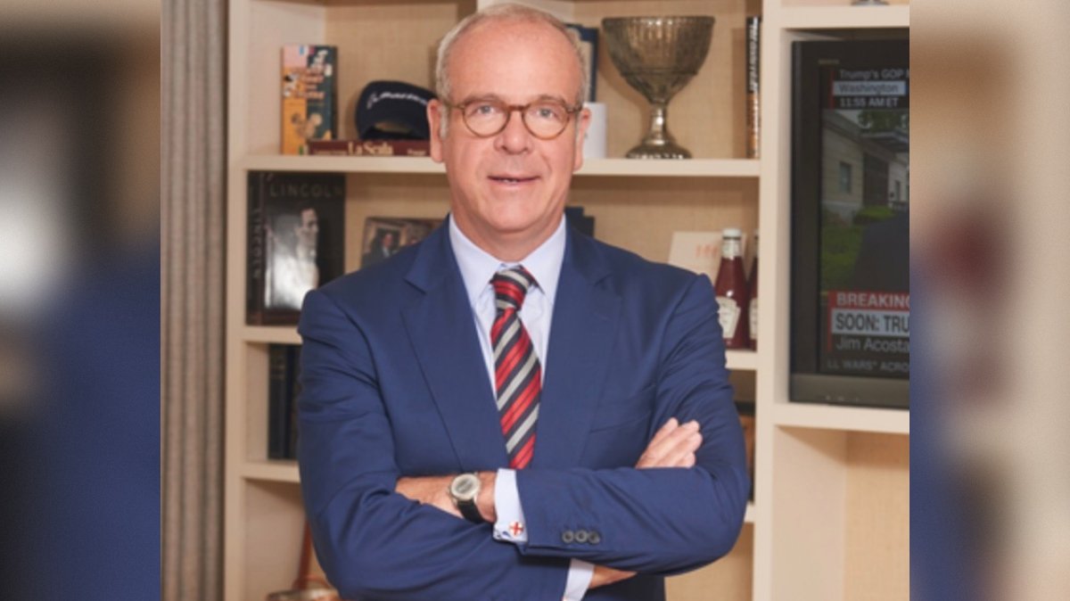 Professional man in a blue suit stands confidently with arms crossed, surrounded by books and a television displaying news content.