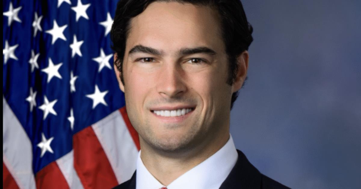 Portrait of a smiling man in a suit with a red tie, standing in front of the American flag, representing a professional political figure.