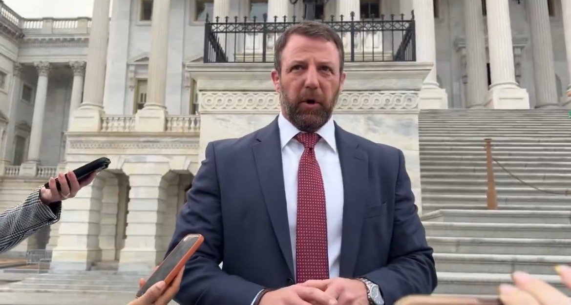 Man in a suit speaking to reporters outside a government building, with several phones pointed towards him for interviews.