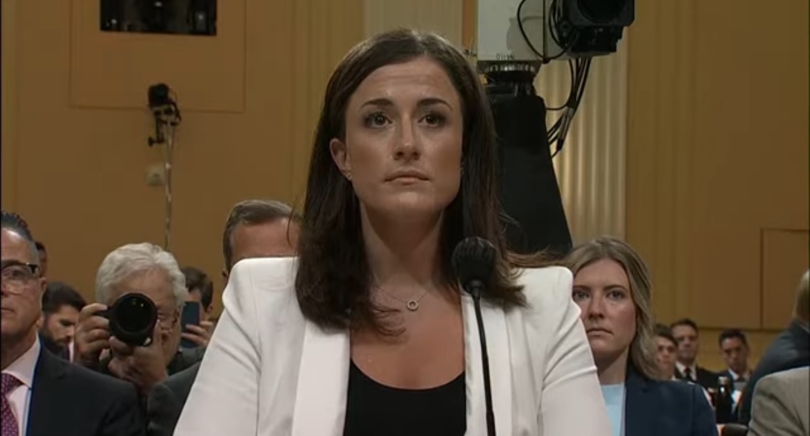 Woman in a white blazer testifying during a congressional hearing, with an audience of attendees and cameras in the background.