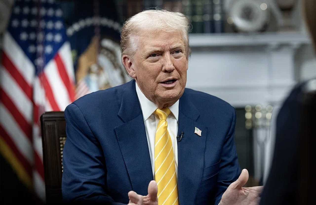 Donald Trump engaged in a discussion, wearing a navy suit and yellow tie, with the American flag and presidential seal in the background.