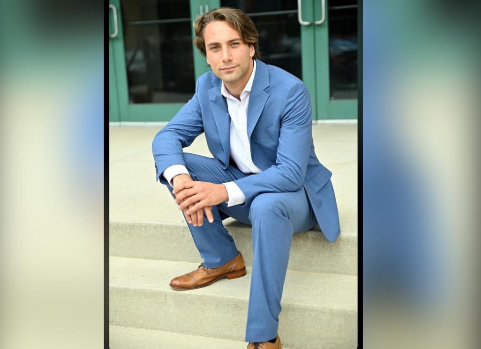Man in a blue suit sitting on steps, looking confidently at the camera with a modern building in the background.