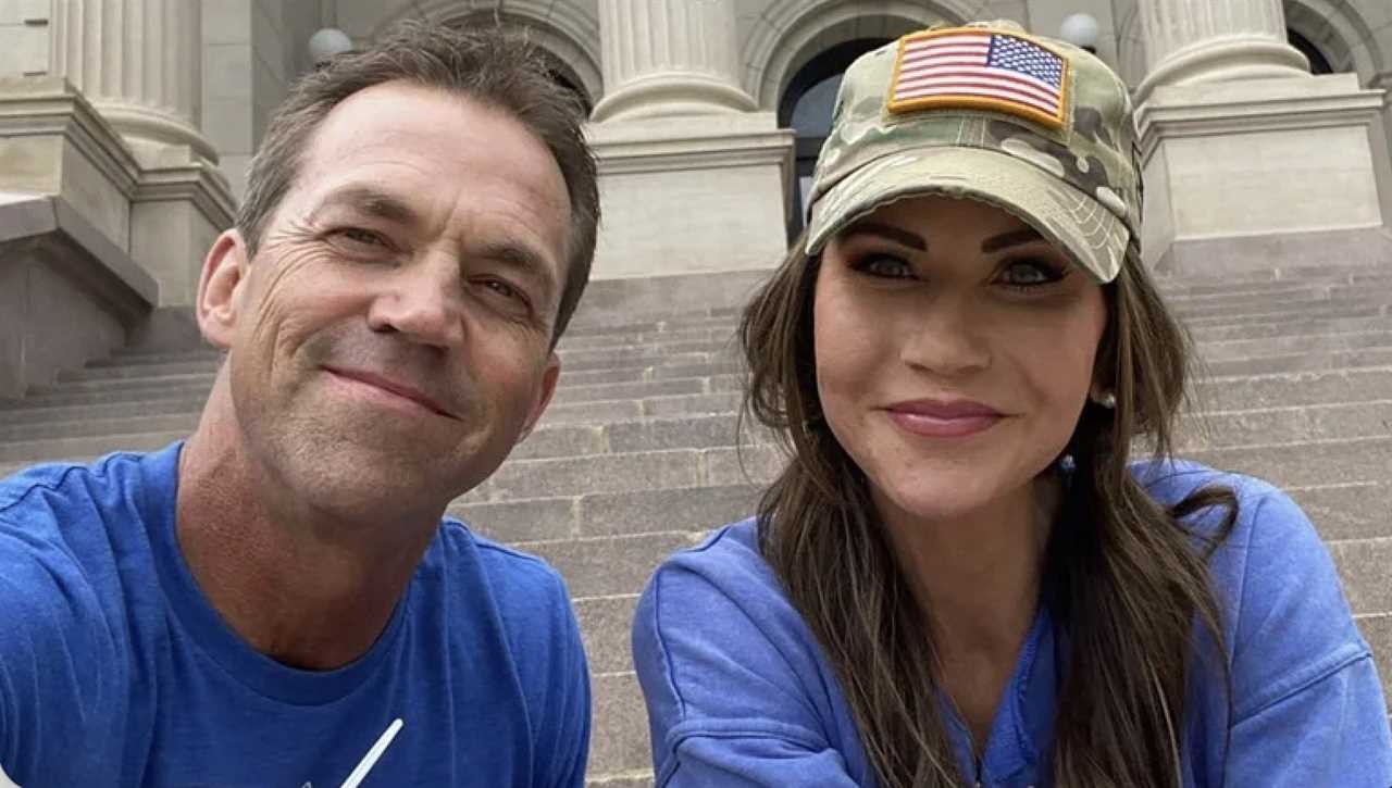 Smiling couple posing for a selfie on outdoor steps, wearing casual blue shirts and a camo hat with an American flag patch.