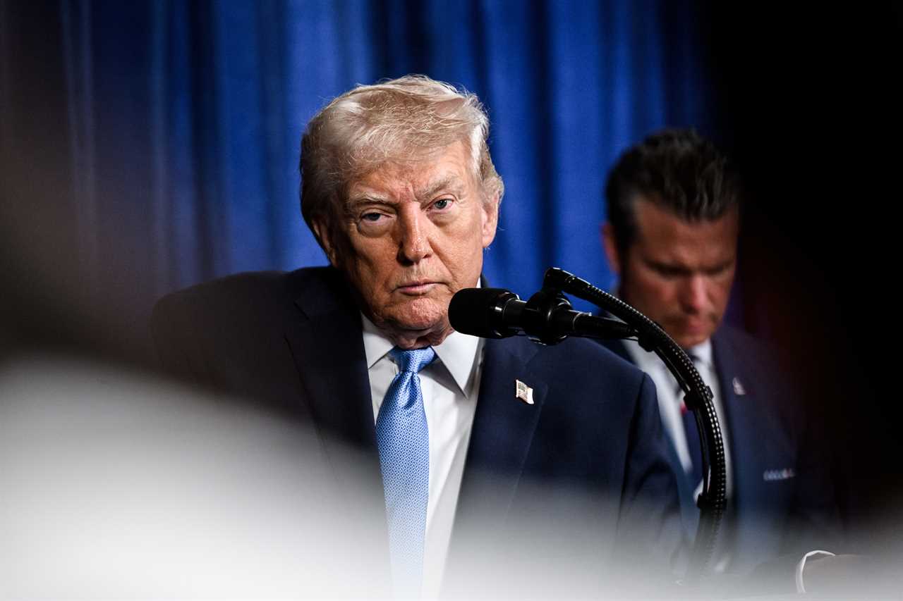 Donald Trump speaking at a podium with a microphone, showing a serious expression during a press event, with a blue backdrop.