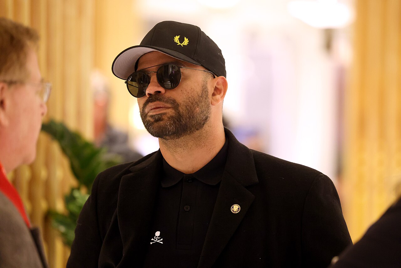 Man wearing a black cap and sunglasses engaged in conversation indoors, showcasing a stylish look with a casual yet sophisticated outfit.