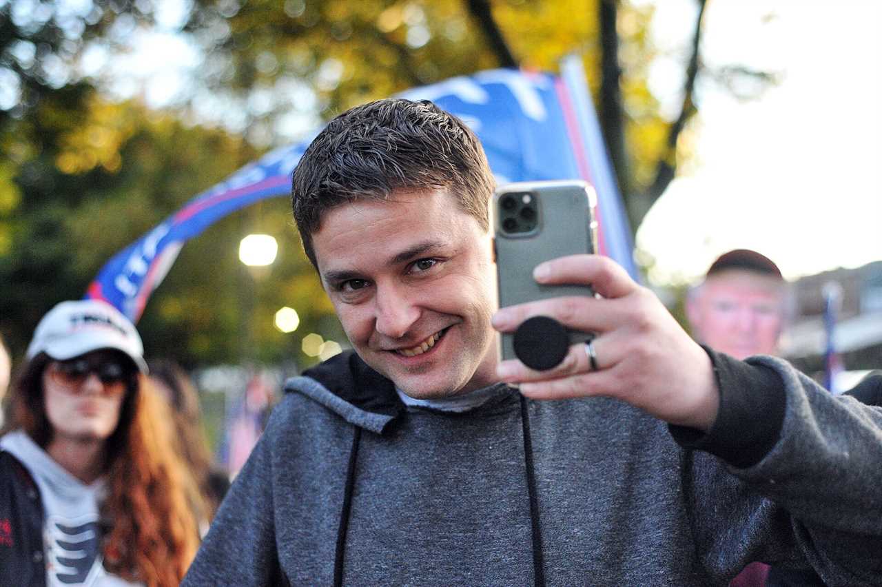 Smiling man in a gray hoodie takes a selfie at an outdoor event with supporters in the background, showcasing a festive atmosphere.