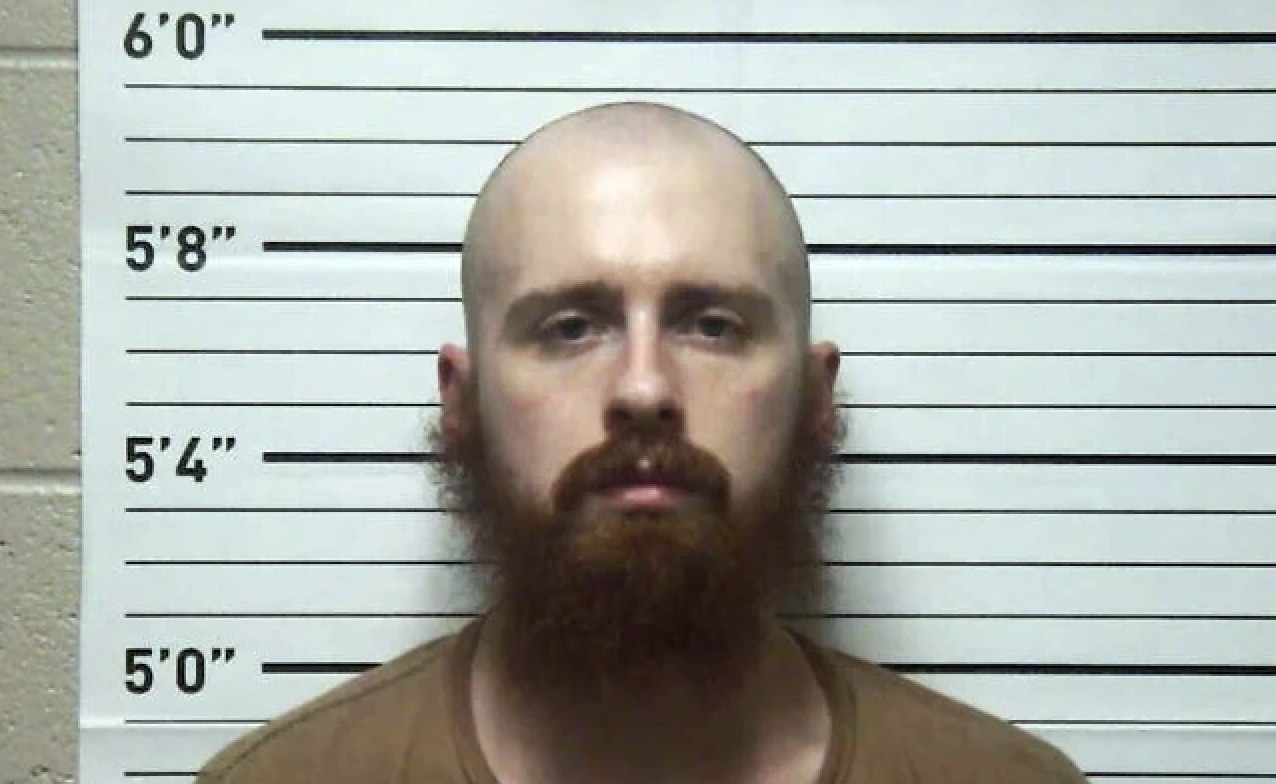 Mugshot of a man with a bald head and red beard, standing against a height chart background.