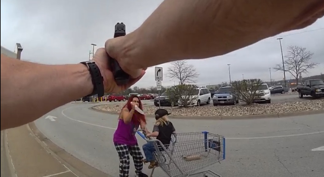 A person points a gun at a woman and child in a shopping cart, capturing a tense moment in a parking lot.
