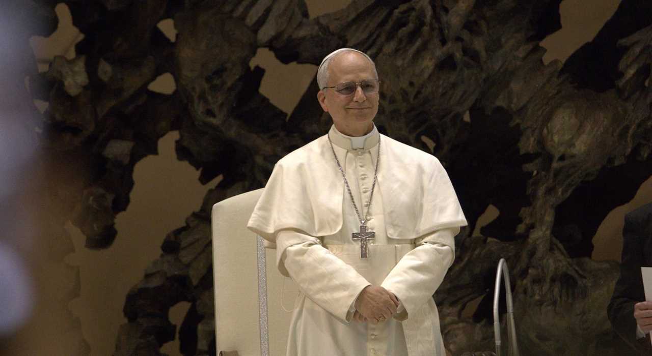 Pope Francis smiles while seated in a formal setting, showcasing his white vestments and distinctive cross against an artistic background.
