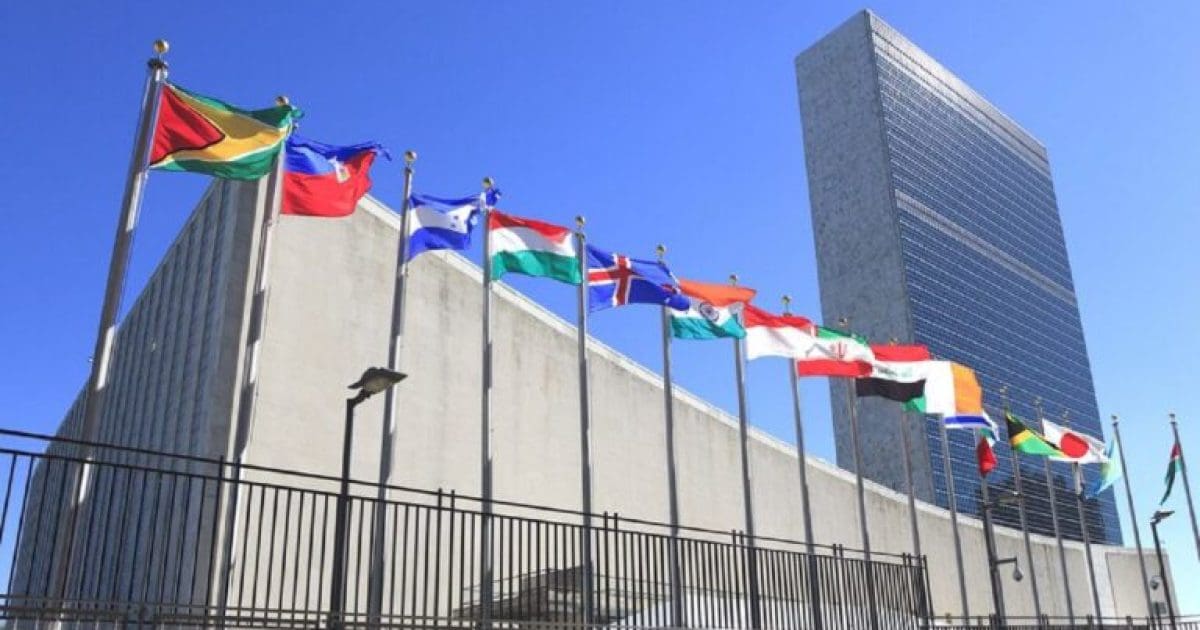 Flags of various nations displayed in front of the United Nations building under a clear blue sky.