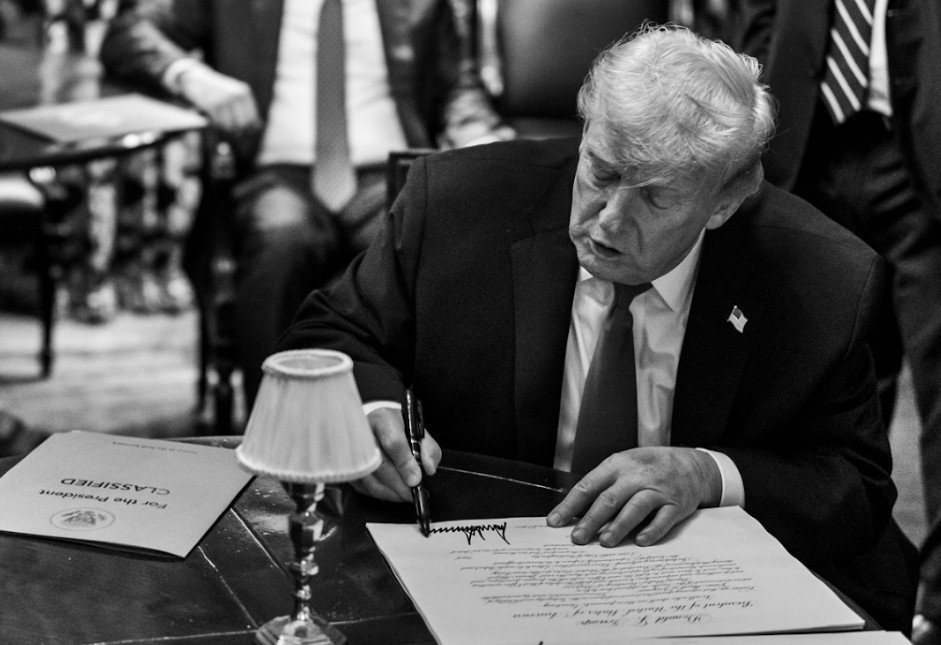 Donald Trump signing a document at a desk, with a lamp and classified papers visible in the background.