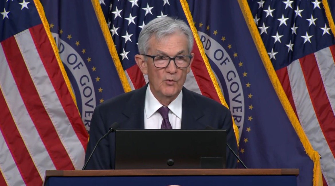 Federal Reserve Chairman delivering a speech at a press conference, with American flags and the Federal Reserve emblem in the background.