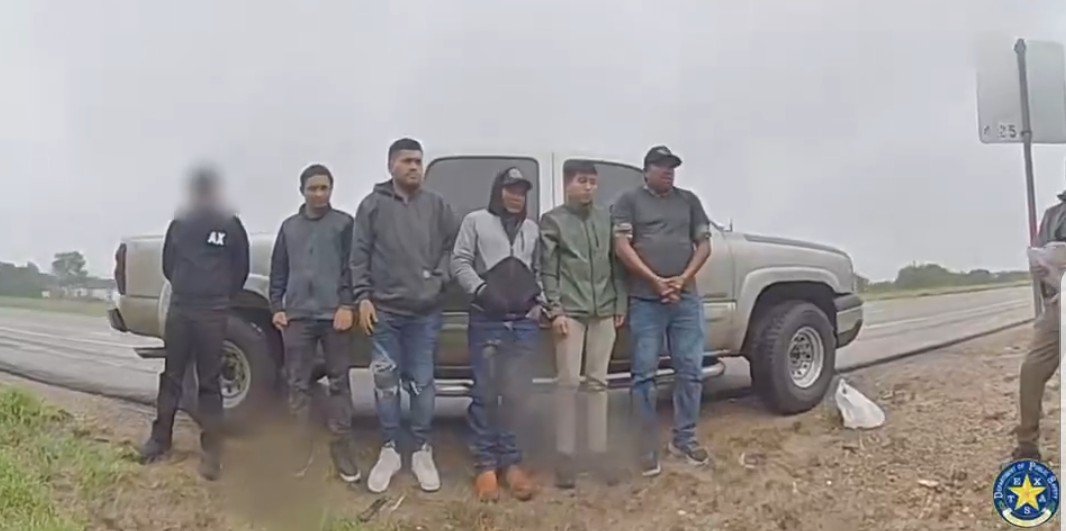 Group of individuals standing by a pickup truck on a roadside, with an officer holding documents nearby in a cloudy, overcast environment.