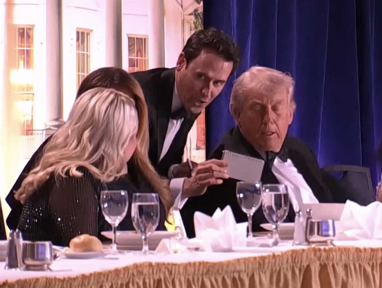 Donald Trump seated at a formal dinner table with guests, engaging with a server who is presenting a note, set against an elegant backdrop.